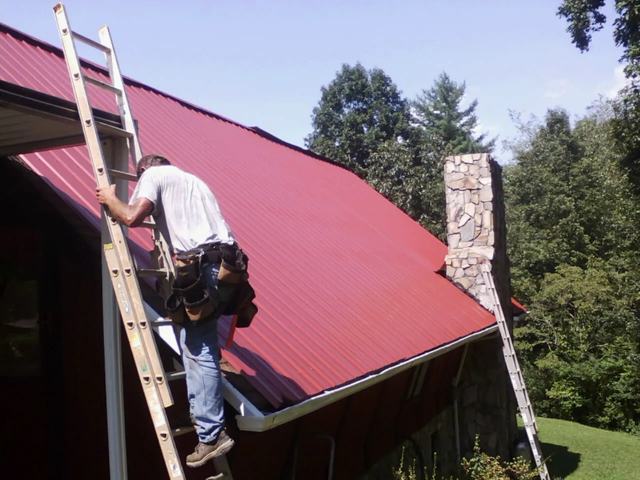 Roofer on a ladder beside a red metal roof and stone chimney — Soffit & Fascia Repair by Roofing Brothers in East Dubuque